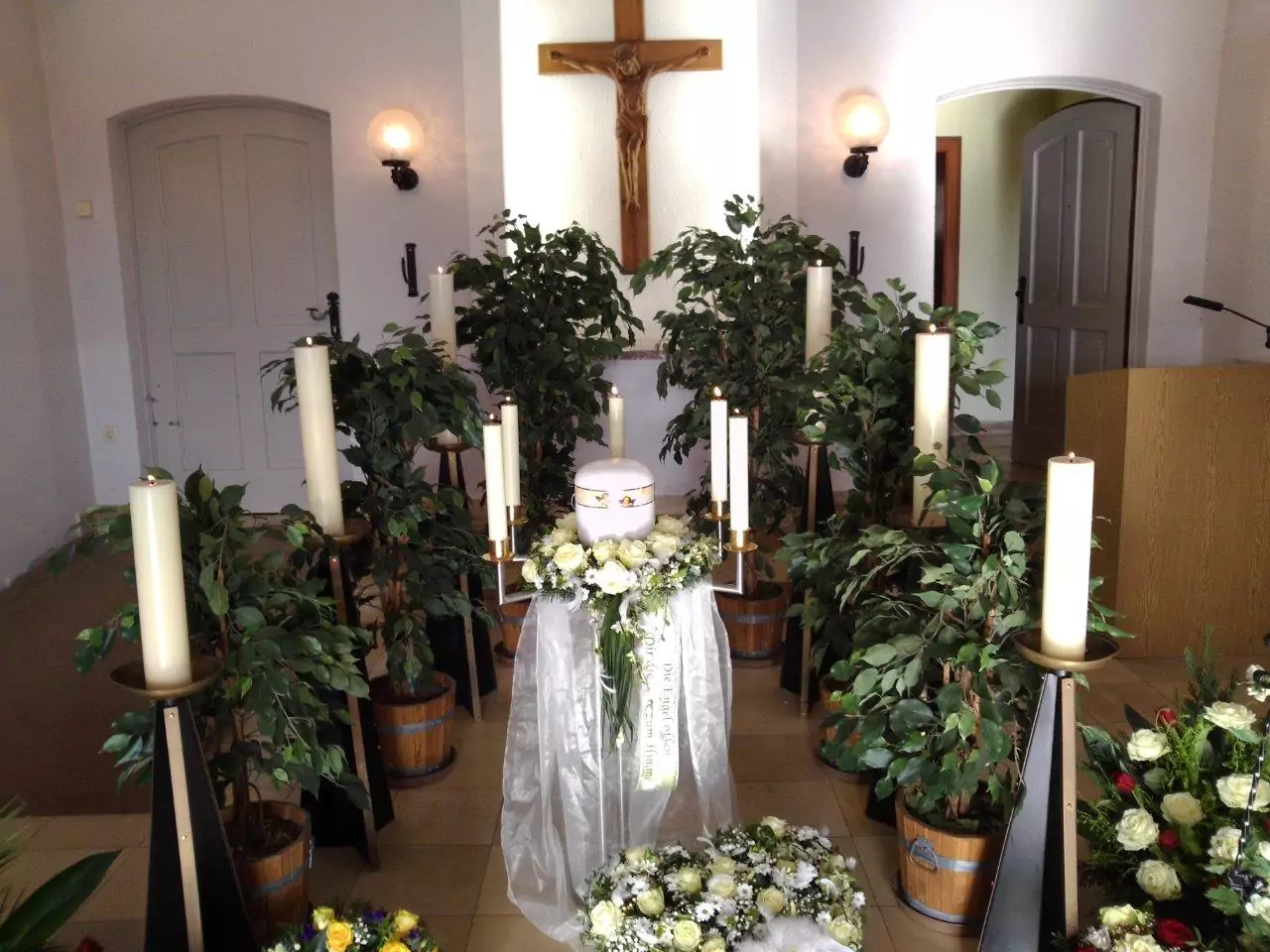 weiße Urne mit Kerzen und Blumenschmuck aus weißen Rosen mit Kreuz im Hintergrund